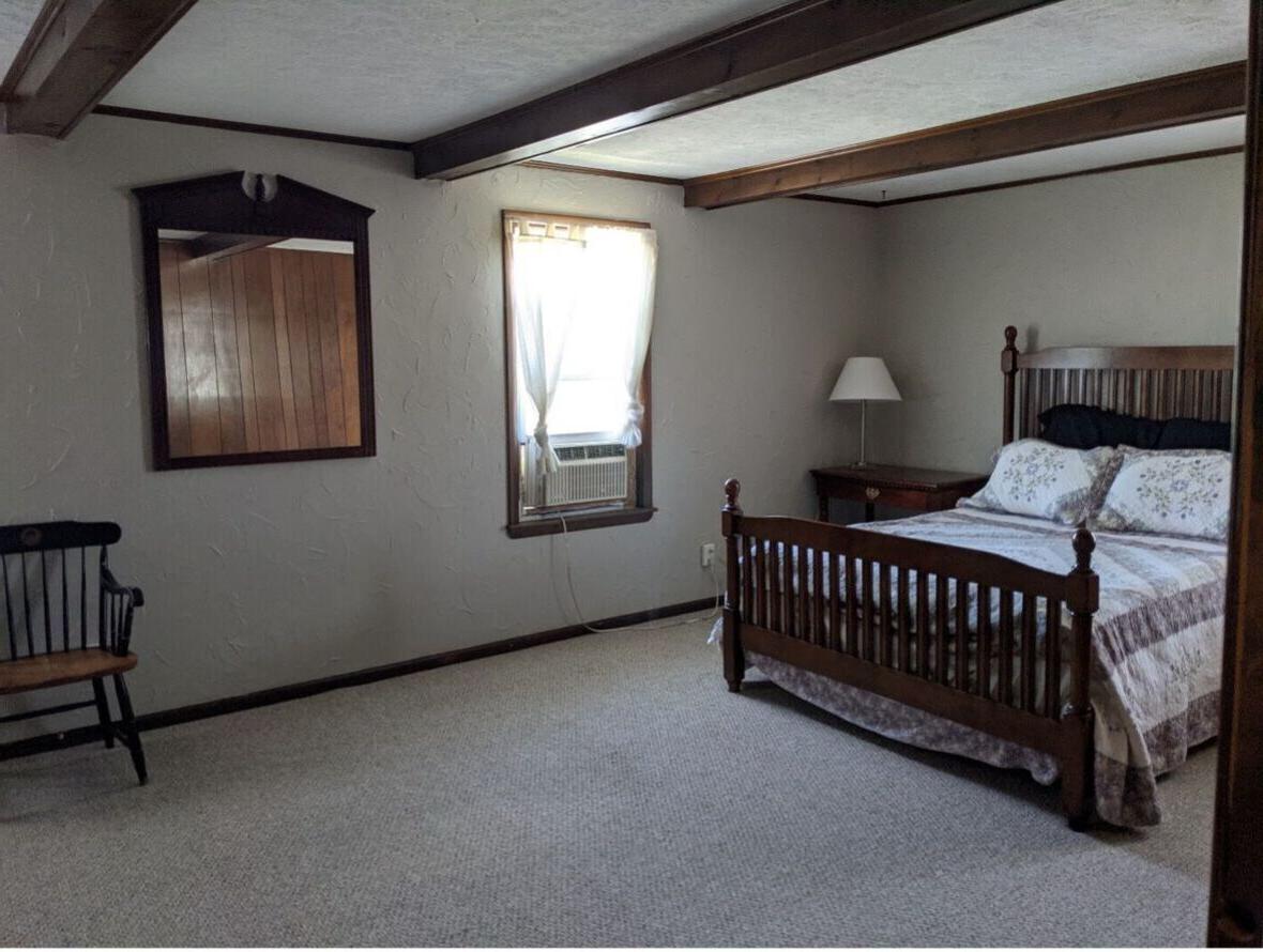 A bedroom in the Alumni House with a full bed in a wooden frame, window with an air conditioning unit, nightstand with lamp, wood-framed mirror, and wooden chair.